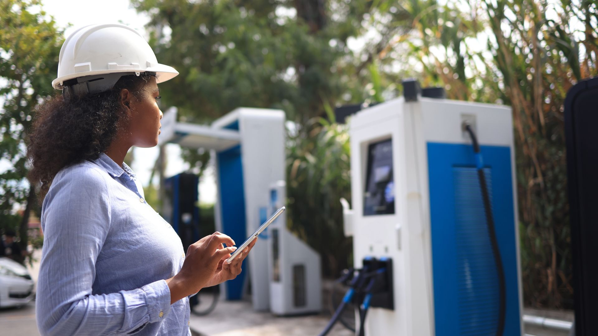Femme portant un casque de chantier utilisant une tablette près de bornes de recharge pour véhicules électriques.
