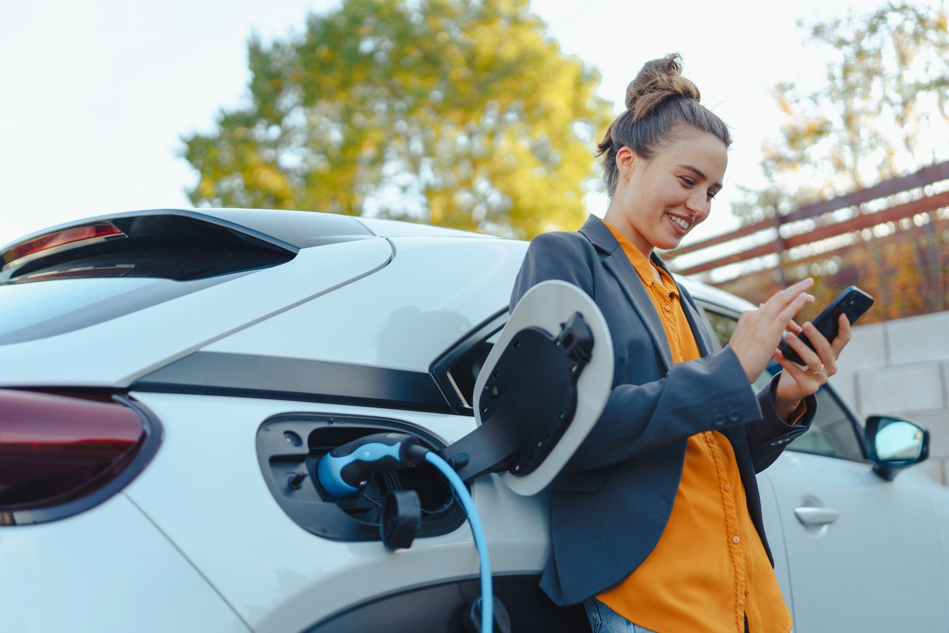 Une femme sourit en utilisant son téléphone à côté d'une voiture électrique branchée et en charge à l'extérieur.