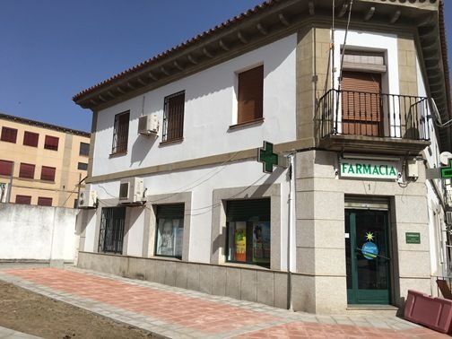 Edificio de farmacia con cartel de cruz verde en una esquina, paredes blancas y puerta marrón.
