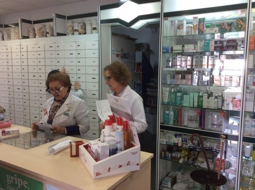 Dos empleados de farmacia examinan papeles tras un mostrador. Estantes con medicamentos detrás.