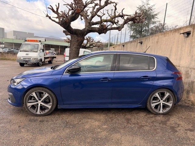 Une Peugeot 308 bleue est garée contre un mur, une dépanneuse est visible en arrière-plan, sous un arbre taillé.