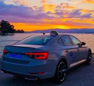 Sed&aacute;n plateado estacionado junto al agua al atardecer, con un cielo colorido y la costa a lo lejos.