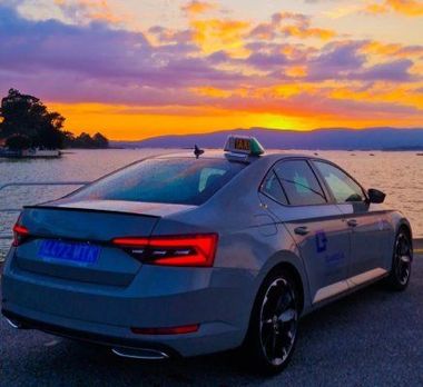 Sed&aacute;n plateado estacionado junto al agua al atardecer, con un cielo colorido y la costa a lo lejos.