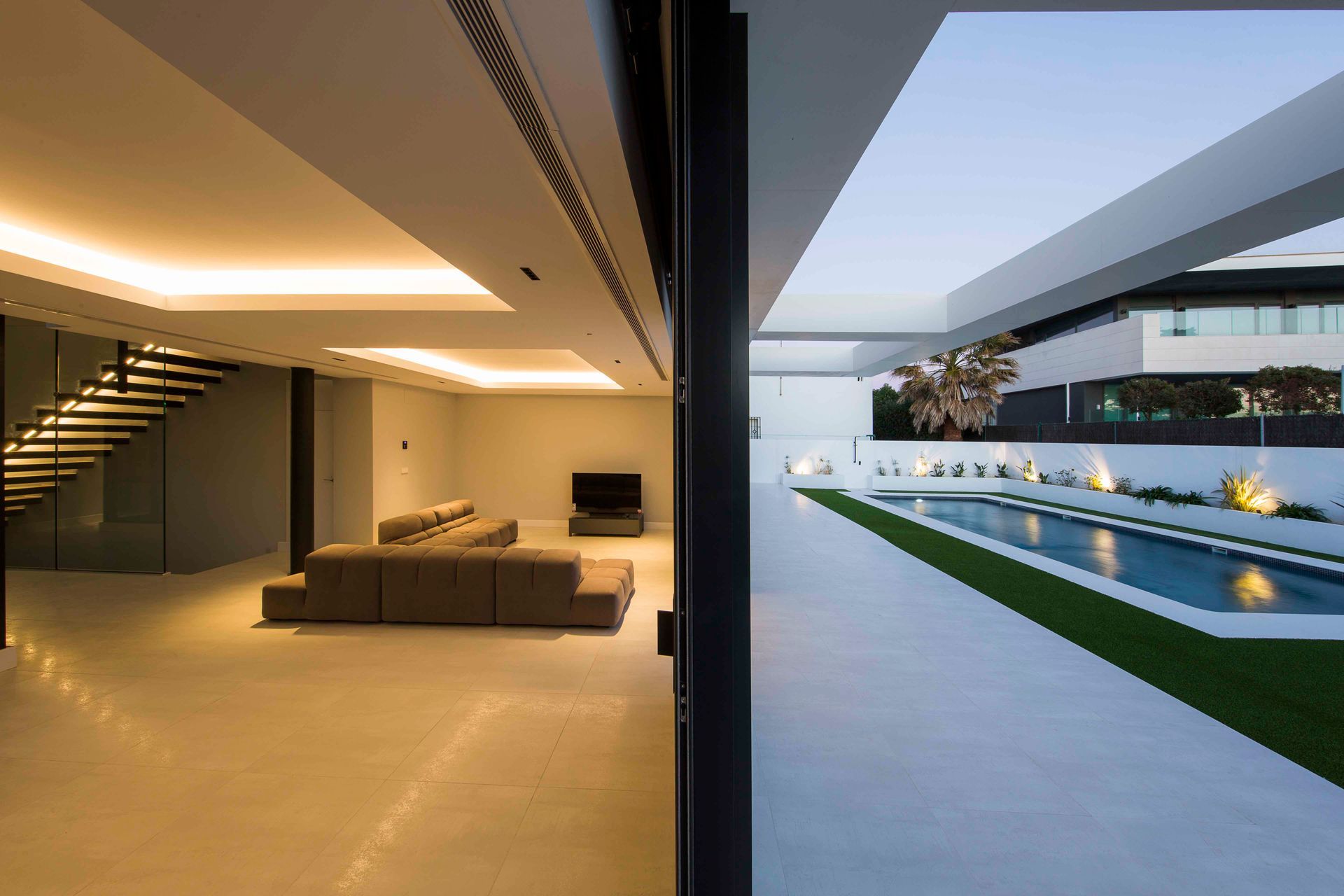 Vista interior y exterior de una casa moderna. Sala de estar con escaleras, sofá y vistas a la piscina.
