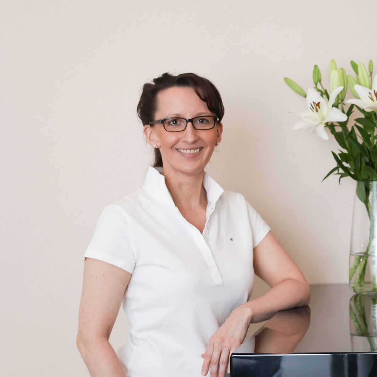 Eine Frau mit Brille und weißem Hemd steht vor einem Schreibtisch.