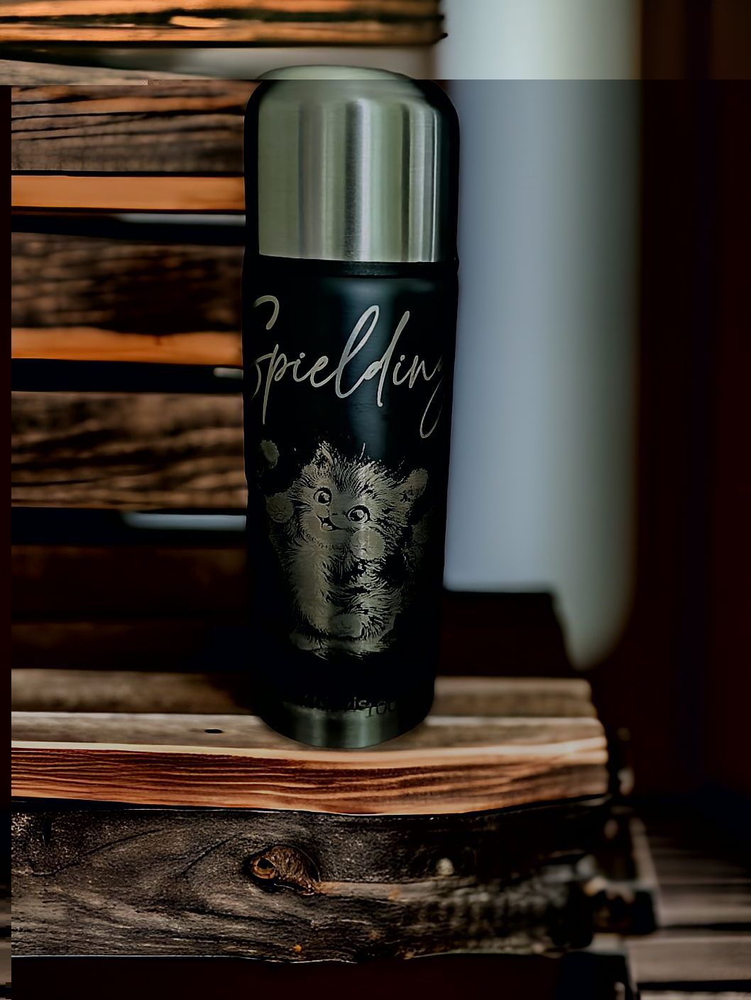 Auf einem Holzregal steht eine schwarze gravierte Thermosflasche.