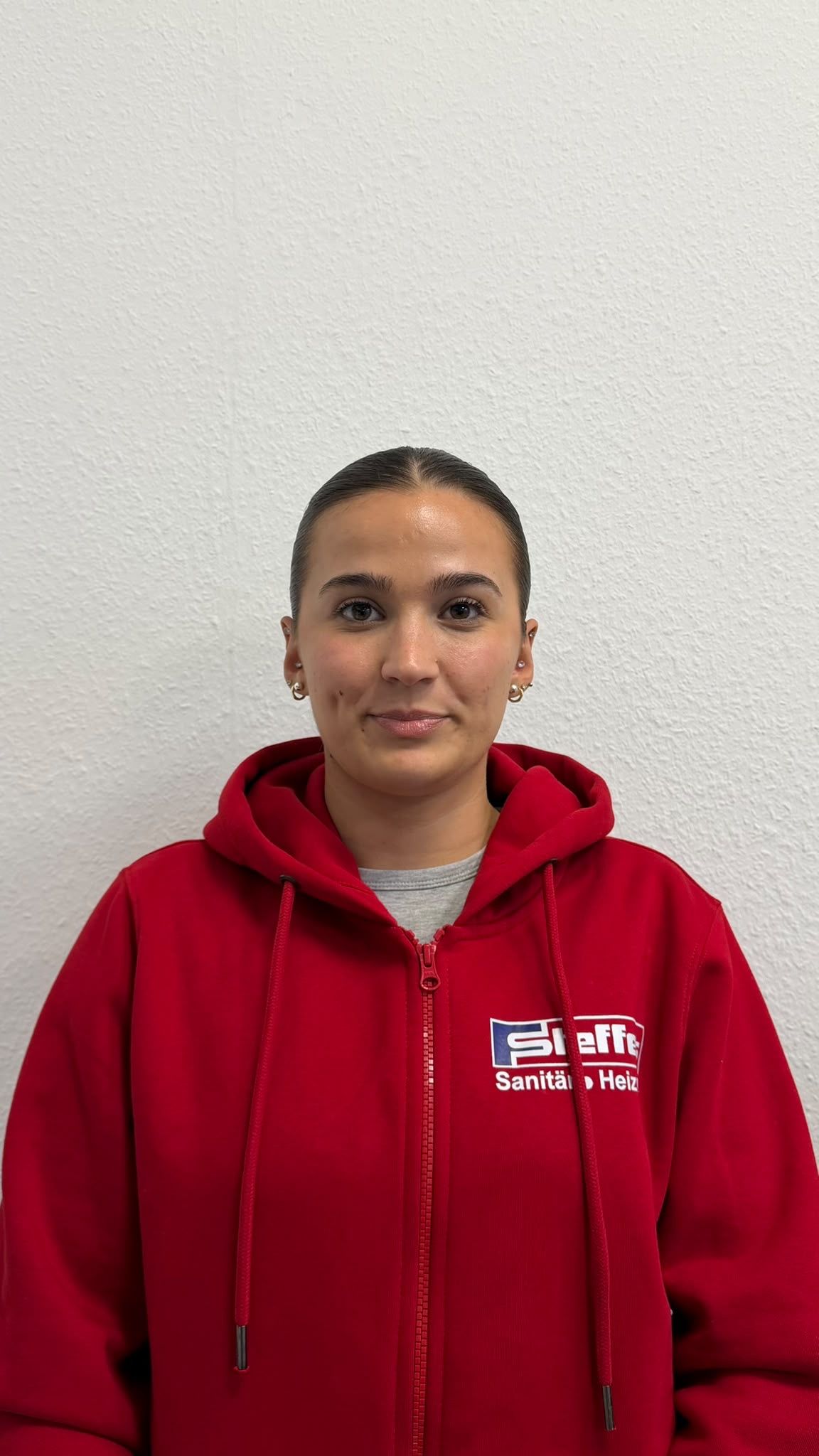 Eine Person in einem roten Kapuzenpullover steht vor einer schlichten Wand und blickt in die Kamera.