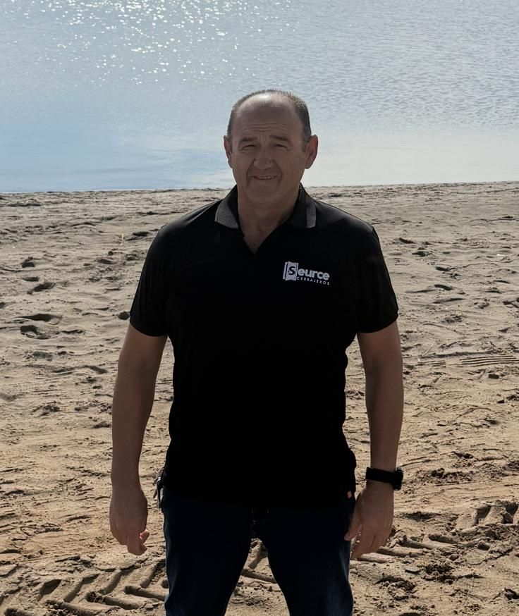 Un hombre de pie en una playa de arena junto a aguas tranquilas, vistiendo una camisa polo negra y sonriendo.