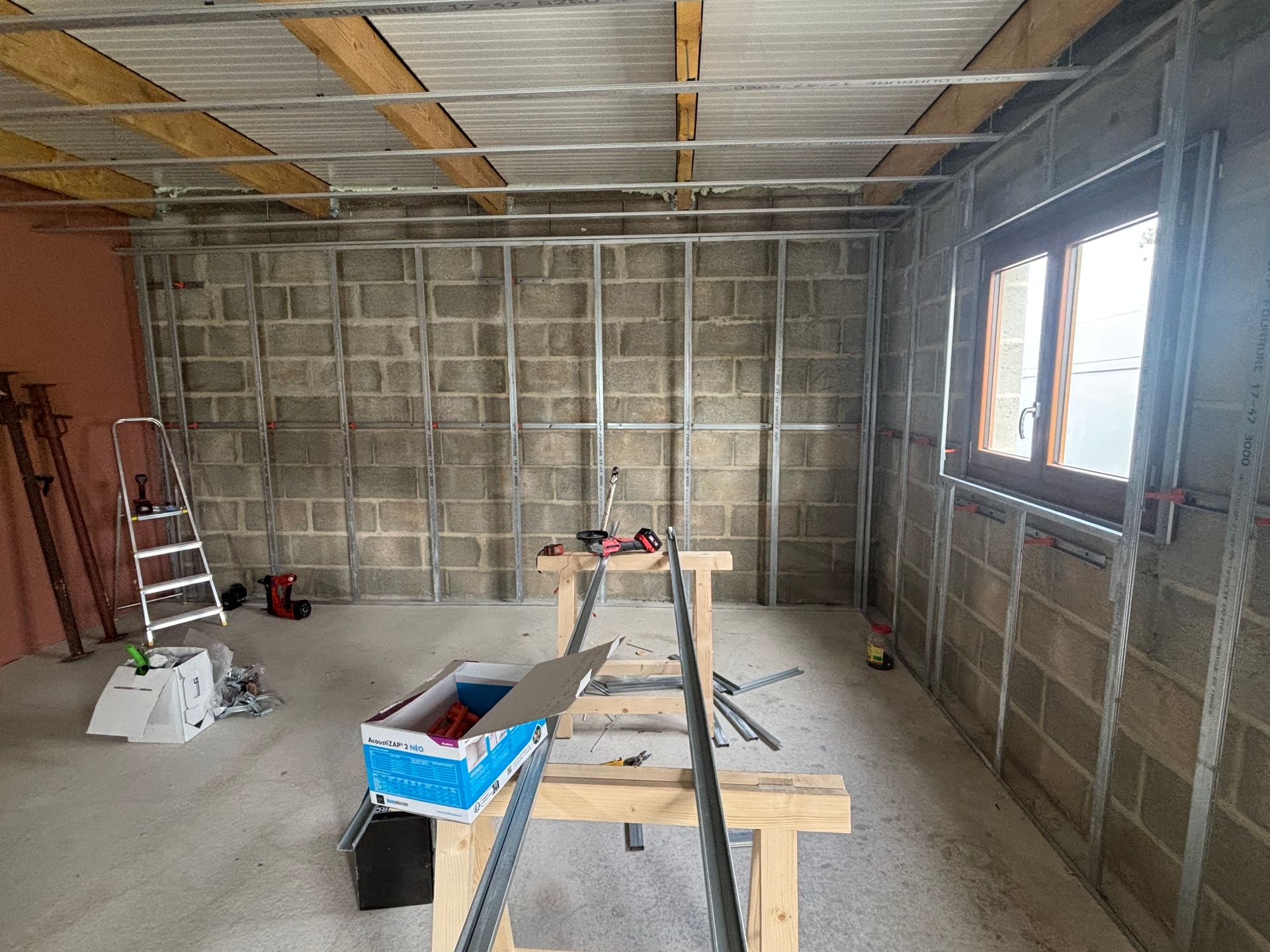 Vue intérieure d'une pièce en construction avec des montants métalliques, des murs en parpaings et une fenêtre.