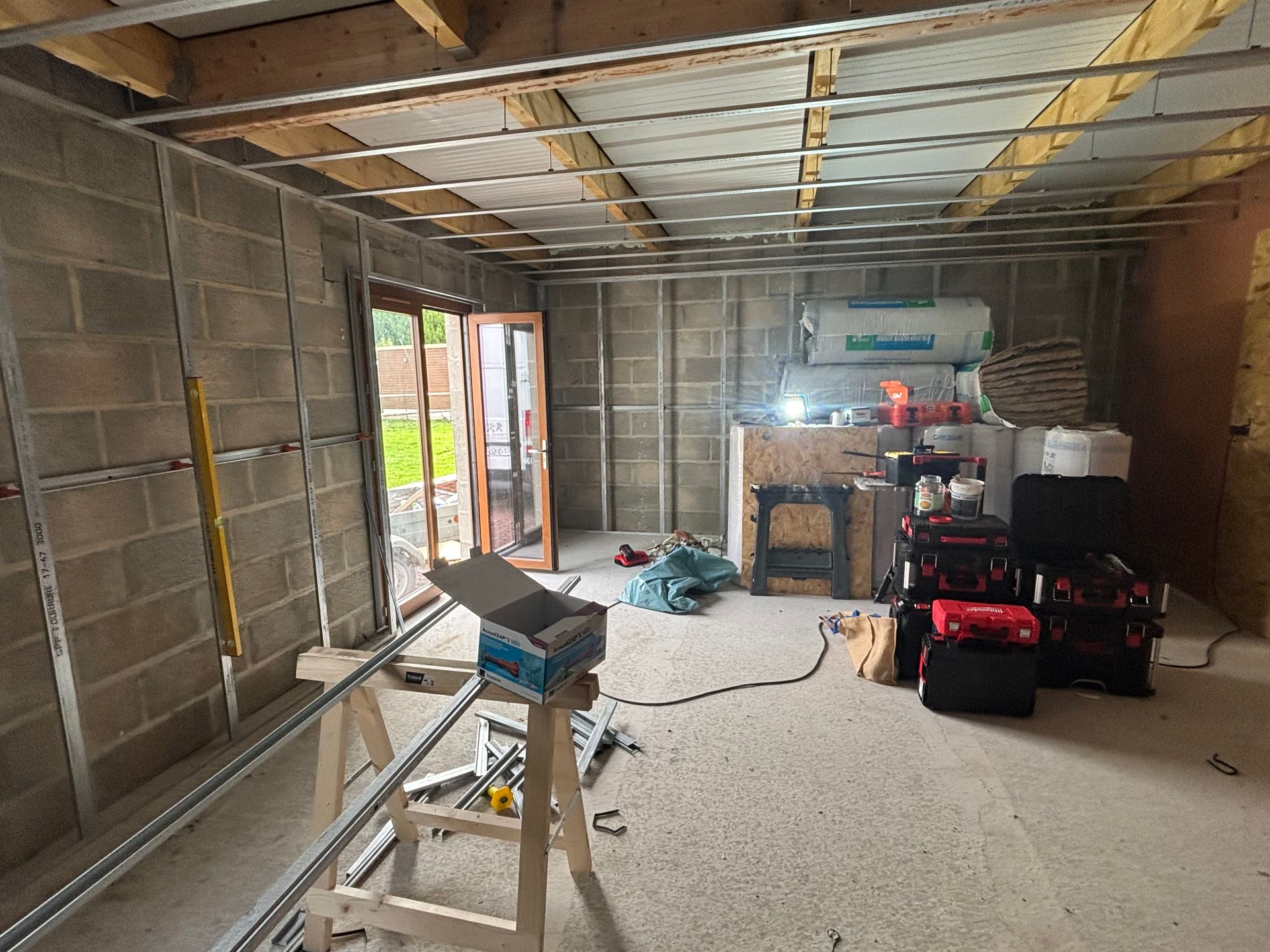 Intérieur d'une pièce en construction avec une structure métallique apparente, des murs en parpaings et une porte ouverte donnant sur une cour arrière.