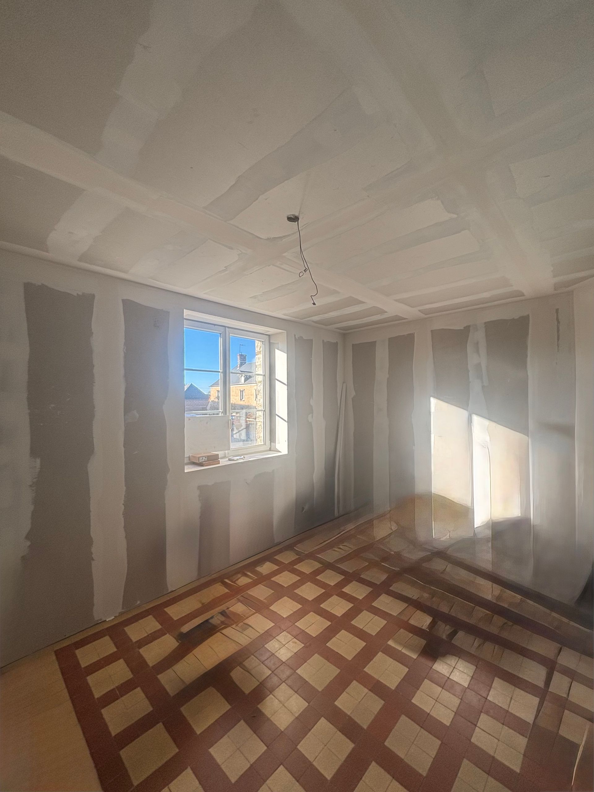 Pièce en travaux avec cloisons sèches apparentes et parquet. La lumière du soleil inonde la pièce par la fenêtre.