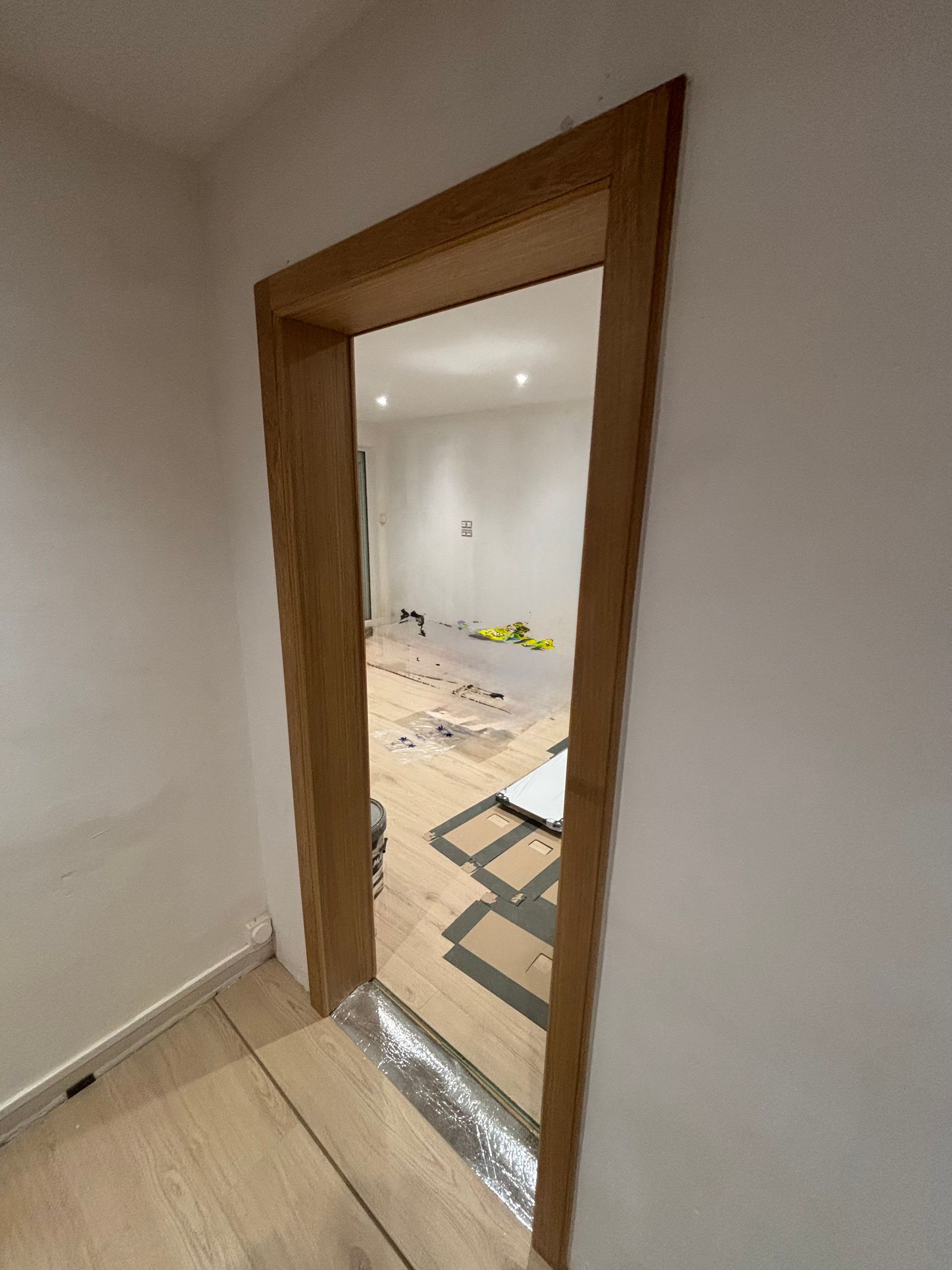 Cadre de porte en bois dans une pièce aux murs blancs, donnant sur une pièce au parquet clair.