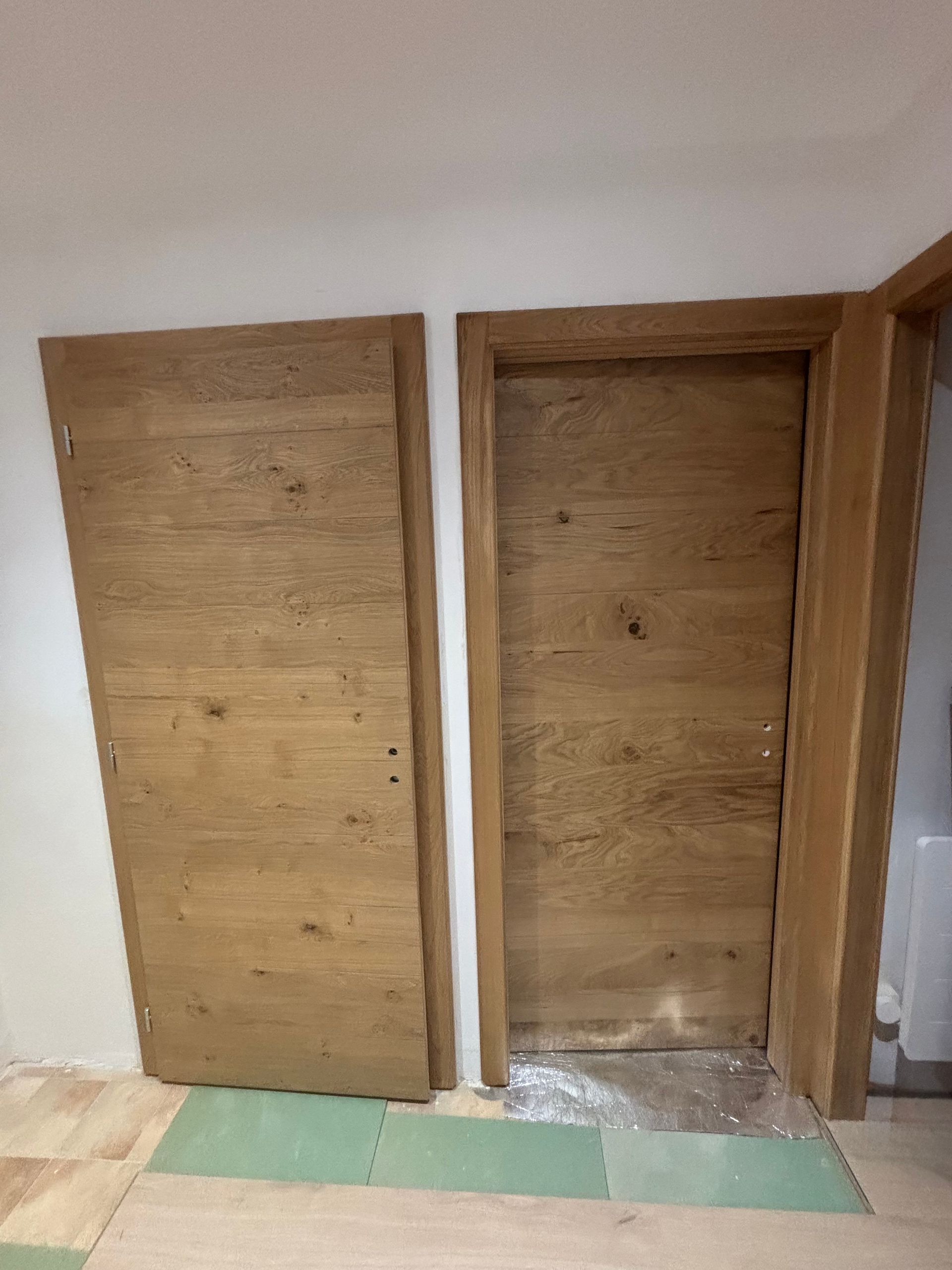 Une porte en bois, contre son cadre, toutes deux avec une finition en bois clair, devant un mur blanc.