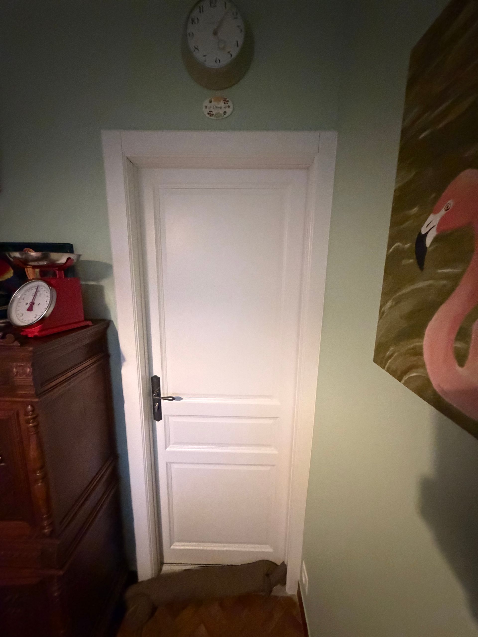 Porte blanche dans une pièce avec une horloge au-dessus, une armoire en bois et un tableau de flamant rose.