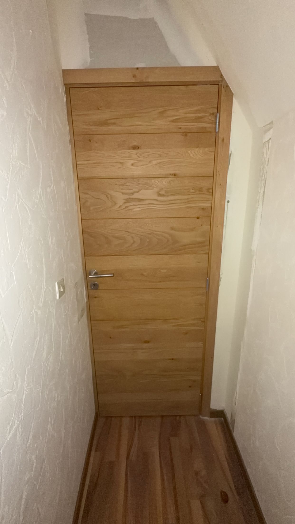 Porte en bois dans un couloir étroit, aux murs et au sol clairs.