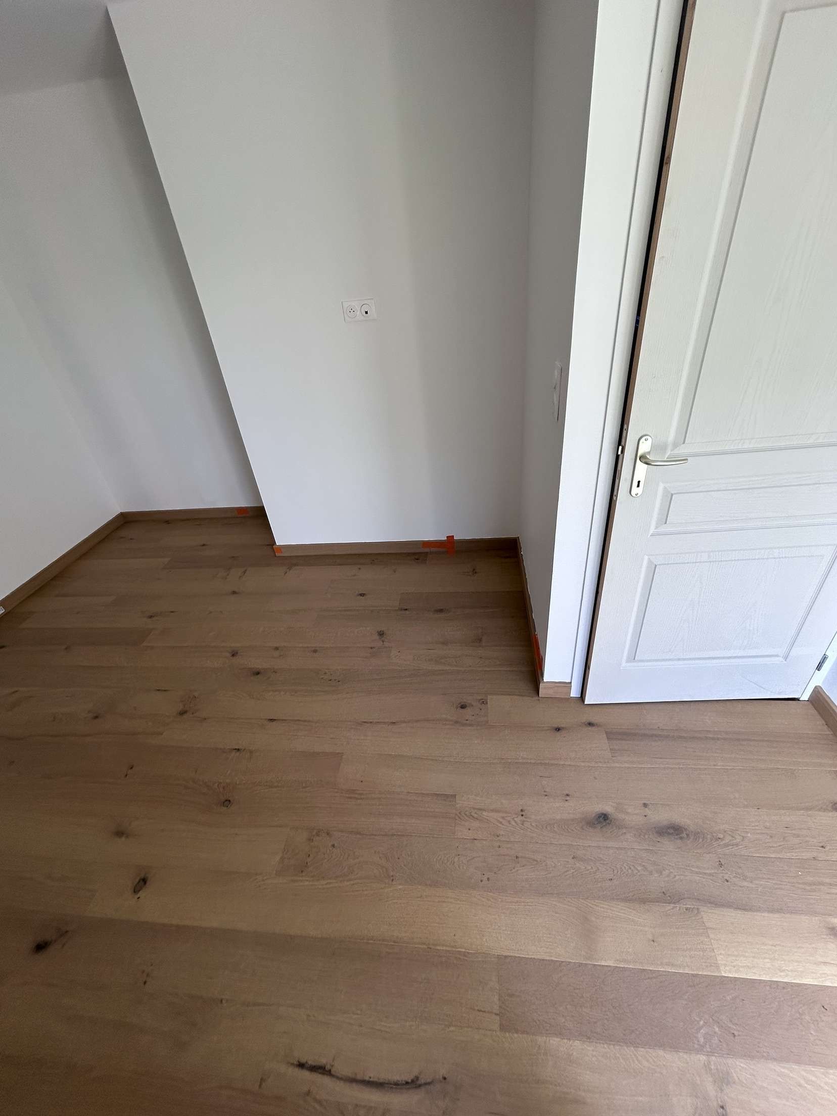 Pièce avec parquet, murs blancs et une porte. Mur en biais à gauche.