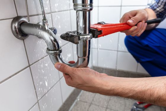 Un plombier resserre un tuyau chromé sous un évier avec une clé rouge alors qu'une fuite d'eau se produit à proximité.