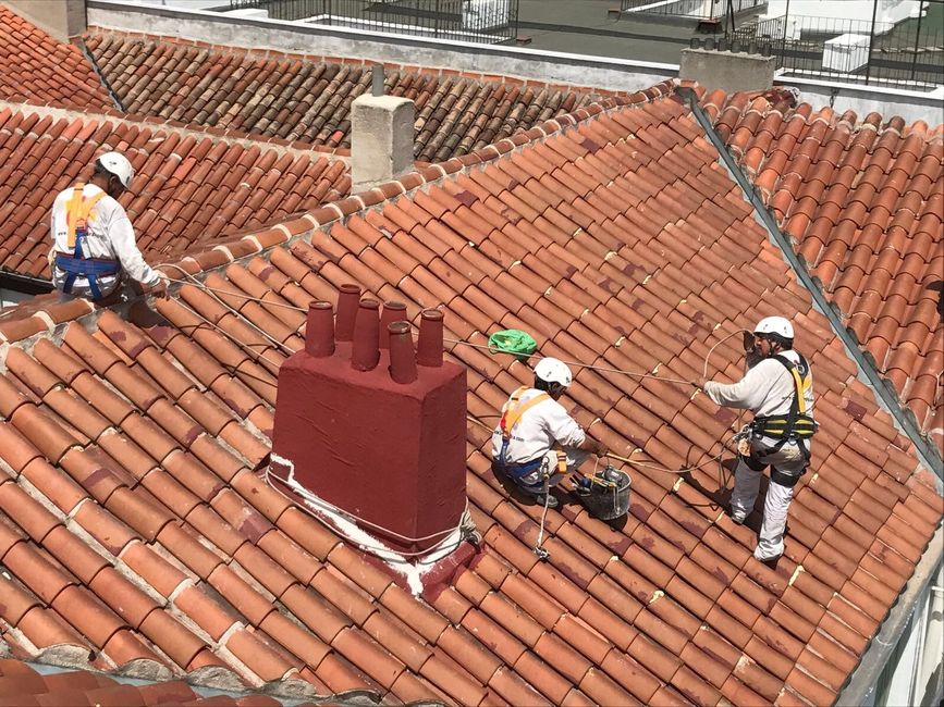 Tres techadores con arneses de seguridad trabajando en un techo de tejas rojas con una chimenea roja.