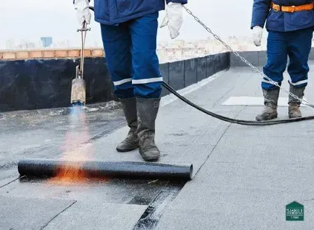 Dos techadores vestidos con ropa de trabajo azul utilizan un soplete para derretir el material del techo en un techo plano.