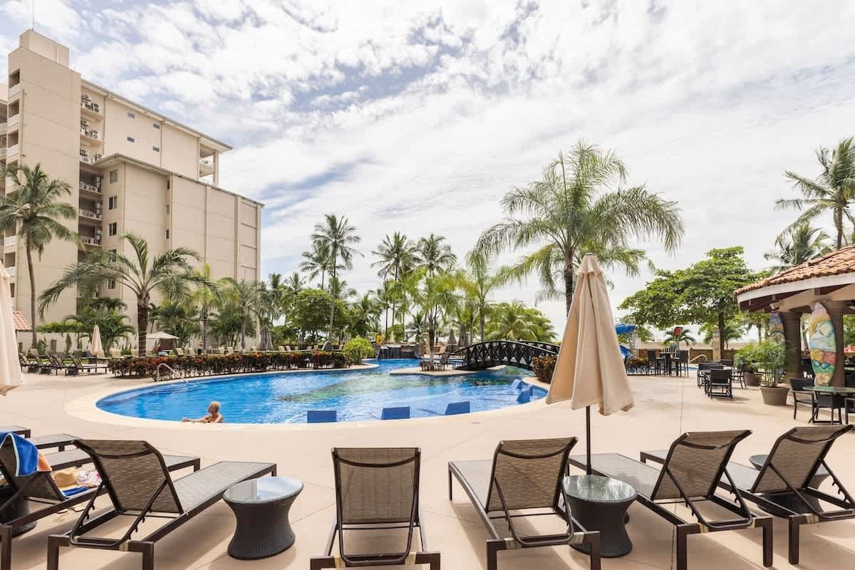 A large swimming pool surrounded by chairs and umbrellas with a building in the background.