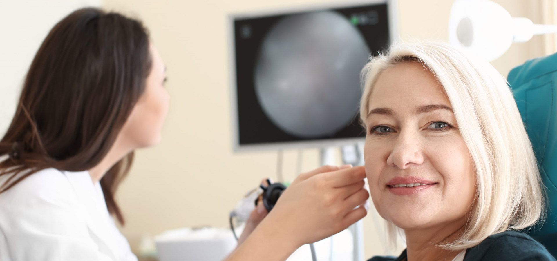 Un profesional examina el oído de un paciente con un otoscopio en un entorno clínico, con un monitor de fondo.