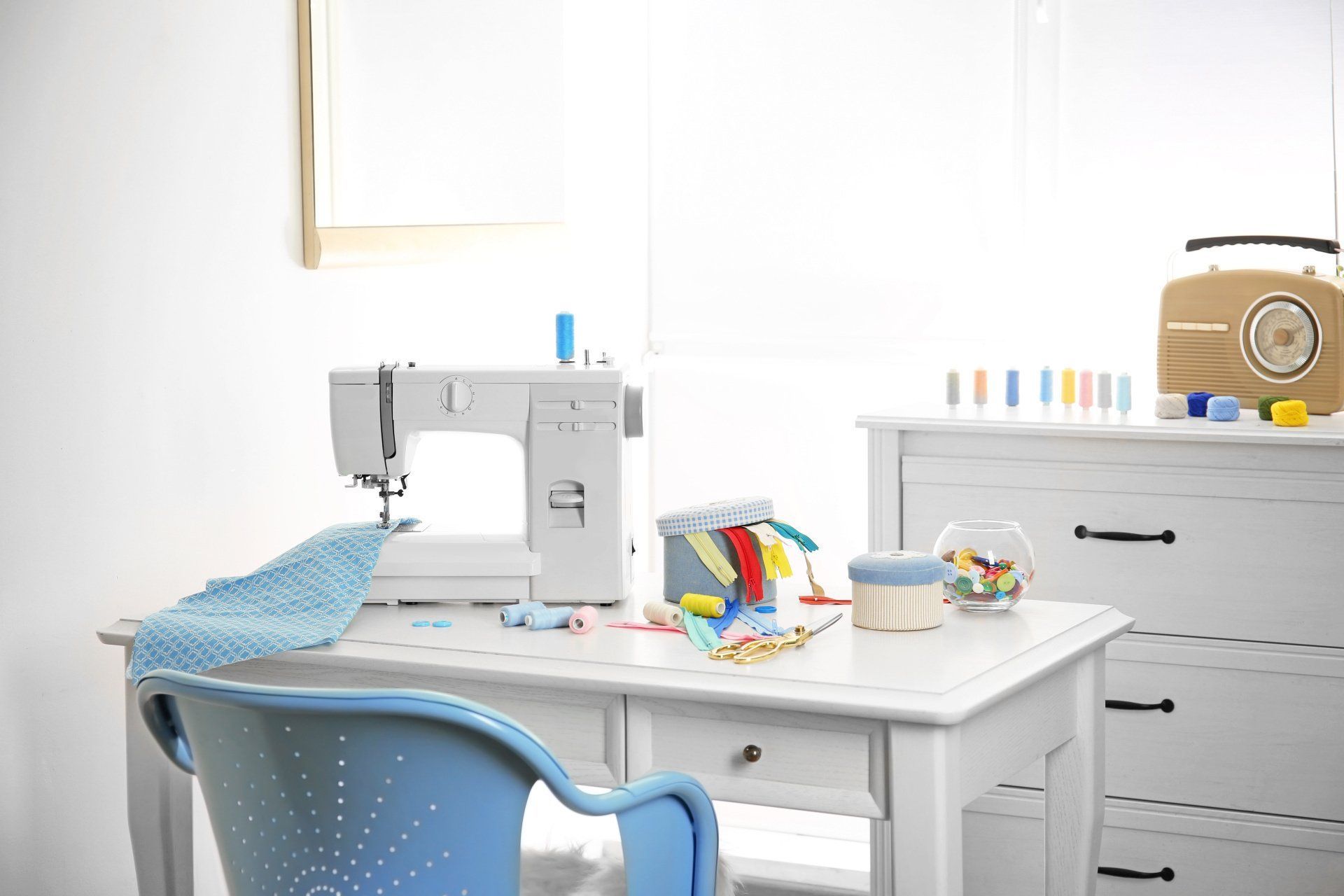 Un espace de couture lumineux avec une machine à coudre blanche, une chaise bleue et des fournitures rangées sur un bureau.