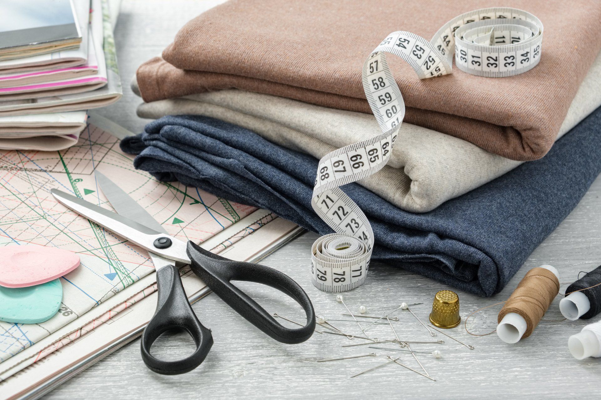 Fournitures de couture avec piles de tissus, mètre ruban, ciseaux, épingles et fil sur une table.