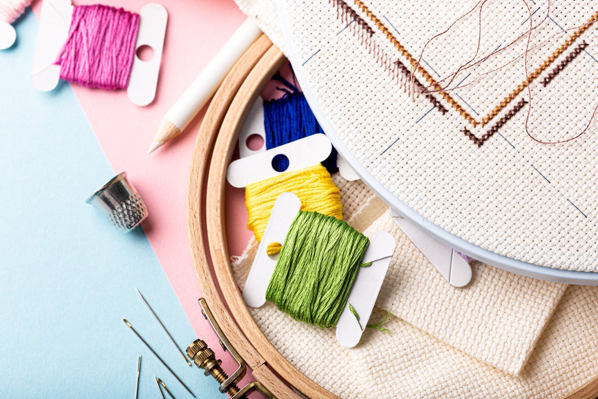 Tambour à broder avec fils colorés, aiguilles et tissu sur un espace de travail rose et bleu.
