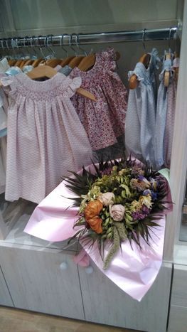 Vestidos de bebé de color rosa colgados en una tienda, con un ramo de flores en primer plano.