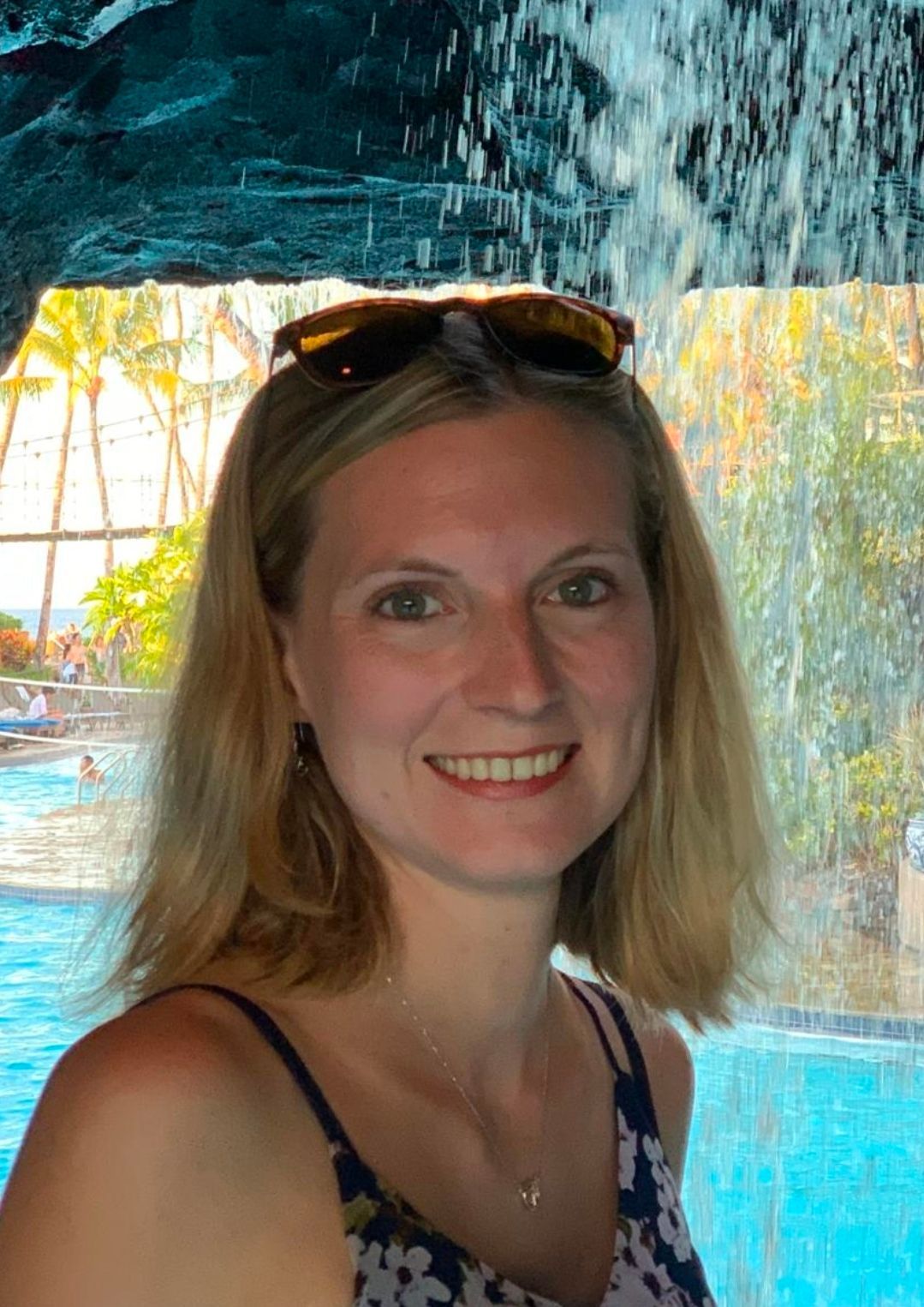 Eine lächelnde Person mit schulterlangem blonden Haar posiert vor einem Wasserfall an einem Resortpool.