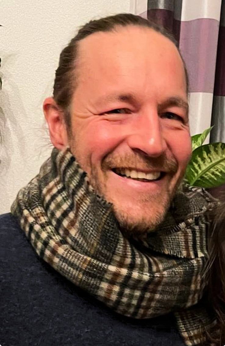 Eine lächelnde Person, die einen dunkelblauen Pullover und einen karierten Schal in Grün-, Braun- und Weißtönen trägt.