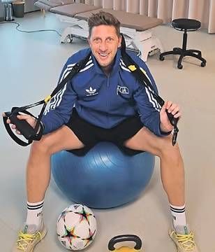 Mann in blauer Adidas-Jacke sitzt auf einem Gymnastikball und benutzt ein Schlingentrainingsgerät mit einem Fussball.