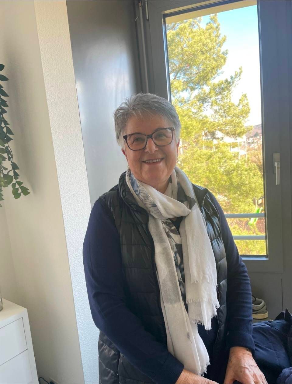 Eine Frau mit Brille und Schal lächelt vor einem Fenster. Sie trägt eine dunkle Weste und einen Pullover.
