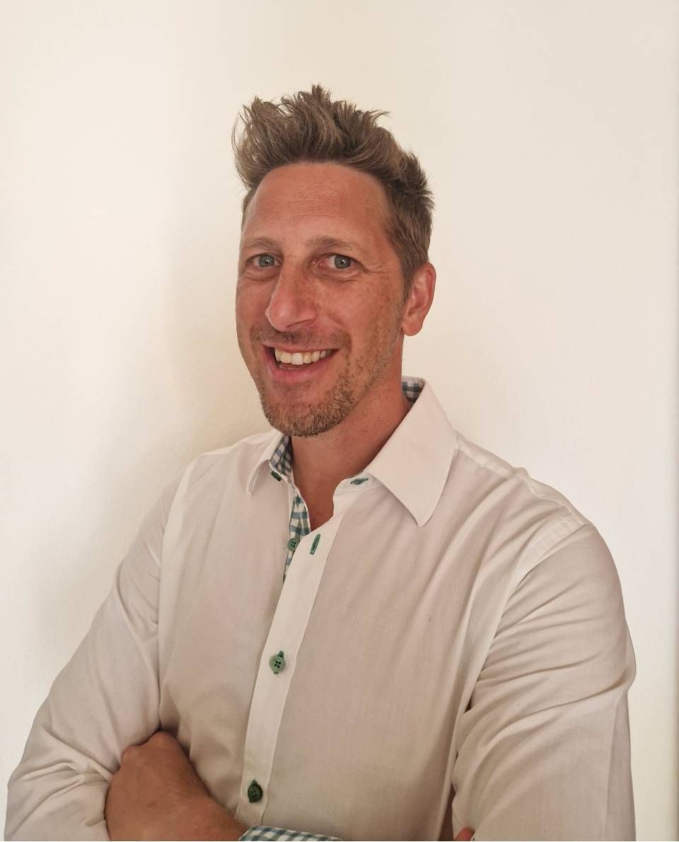 Ein Mann mit verschränkten Armen lächelt; er trägt ein weisses Button-Down-Hemd mit grün gemustertem Kragen.