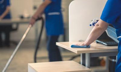 Eine Person, die blaue Handschuhe trägt, reinigt ein Fenster mit einer Sprühflasche und einem gelben Tuch.