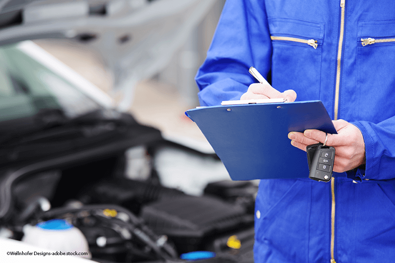 Ein Mechaniker in blauer Arbeitskleidung schreibt auf einem Klemmbrett, während er vor dem geöffneten Motorraum eines Autos die Autoschlüssel in der Hand hält.