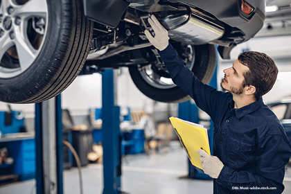 Ein Techniker in Arbeitskleidung und mit Handschuhen inspiziert die Unterseite eines angehobenen Autos in einer Garage, während er eine gelbe Mappe in der Hand hält.