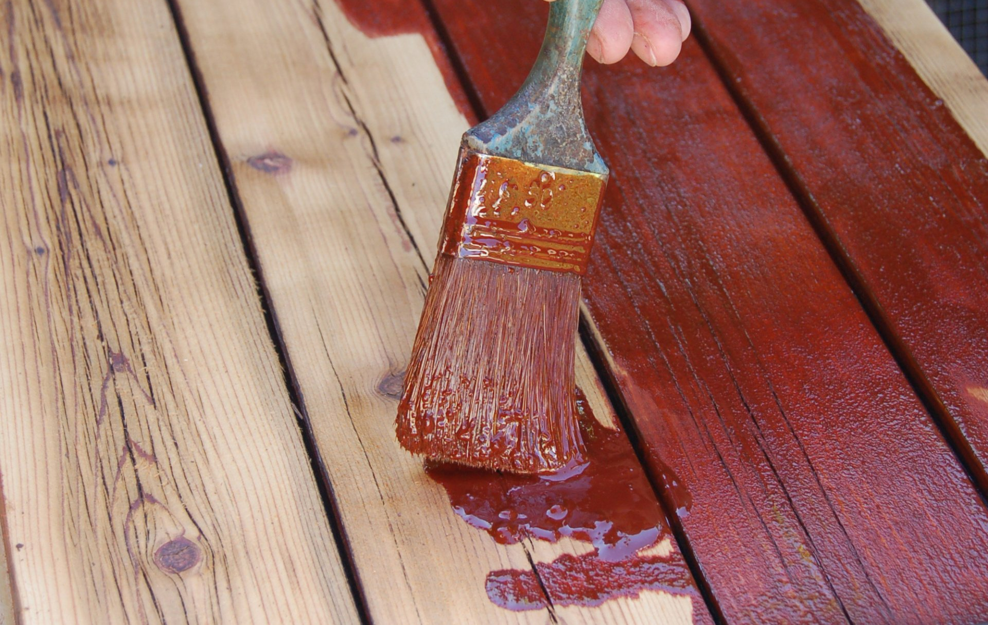 Une main utilisant un pinceau pour appliquer une teinture brun rougeâtre sur des planches de bois.