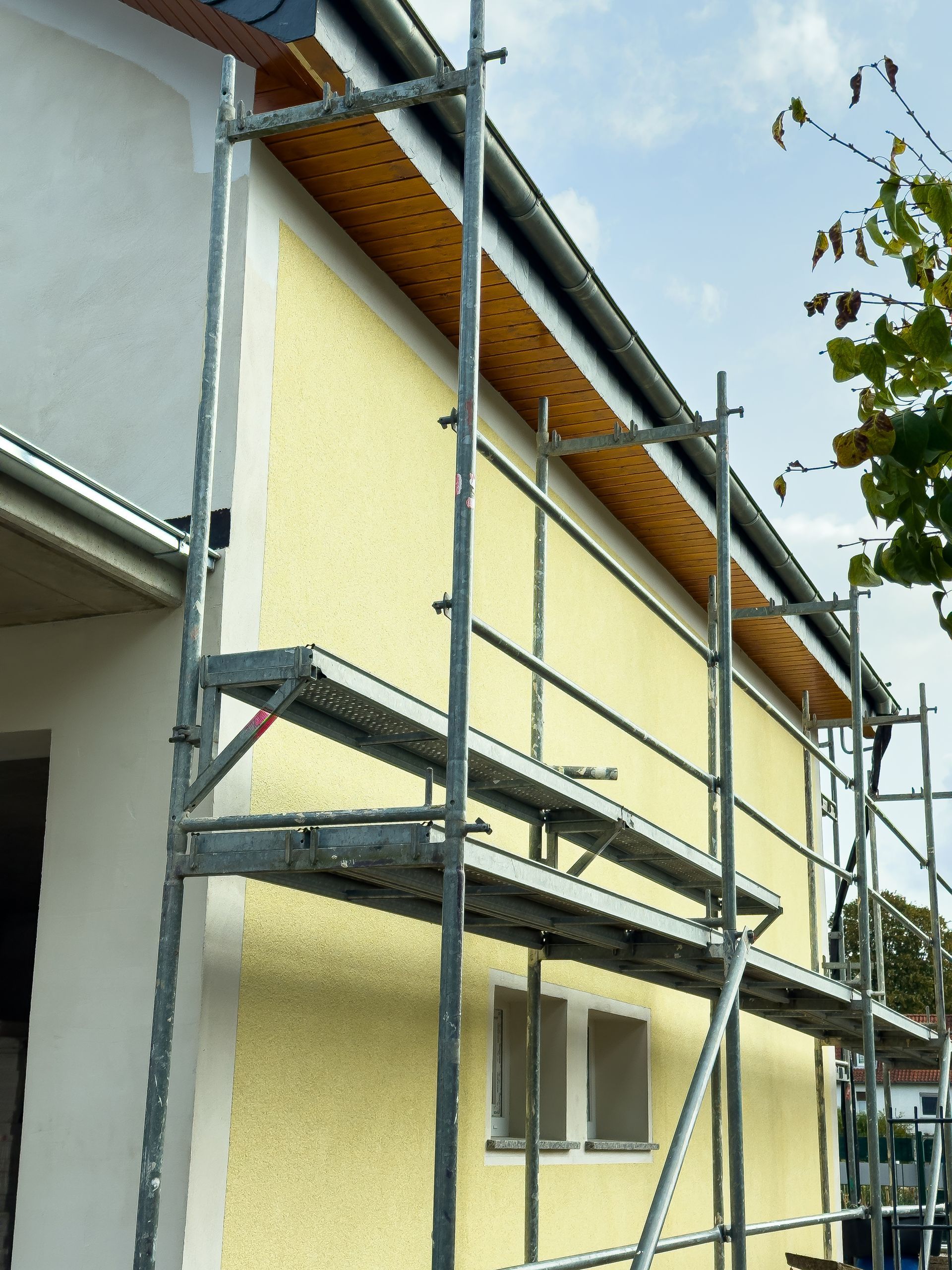 Échafaudage contre un bâtiment peint en jaune avec des avant-toits bruns ; une petite fenêtre est visible.