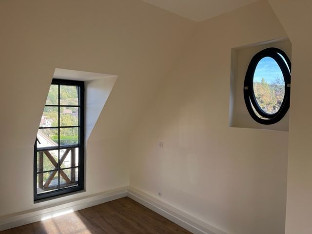 Angle intérieur d'une pièce avec deux fenêtres. L'une est rectangulaire avec un encadrement noir, l'autre ovale avec un encadrement noir. Murs blancs, parquet.