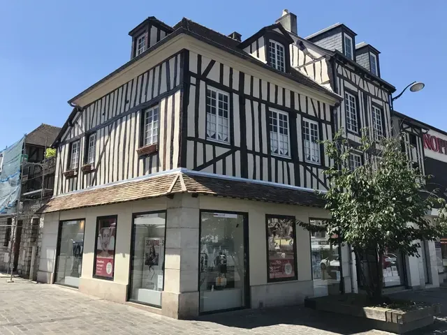 Bâtiment à colombages avec façade de magasin, poutres blanches et noires, toit en pente et arbre devant, par une journée ensoleillée.