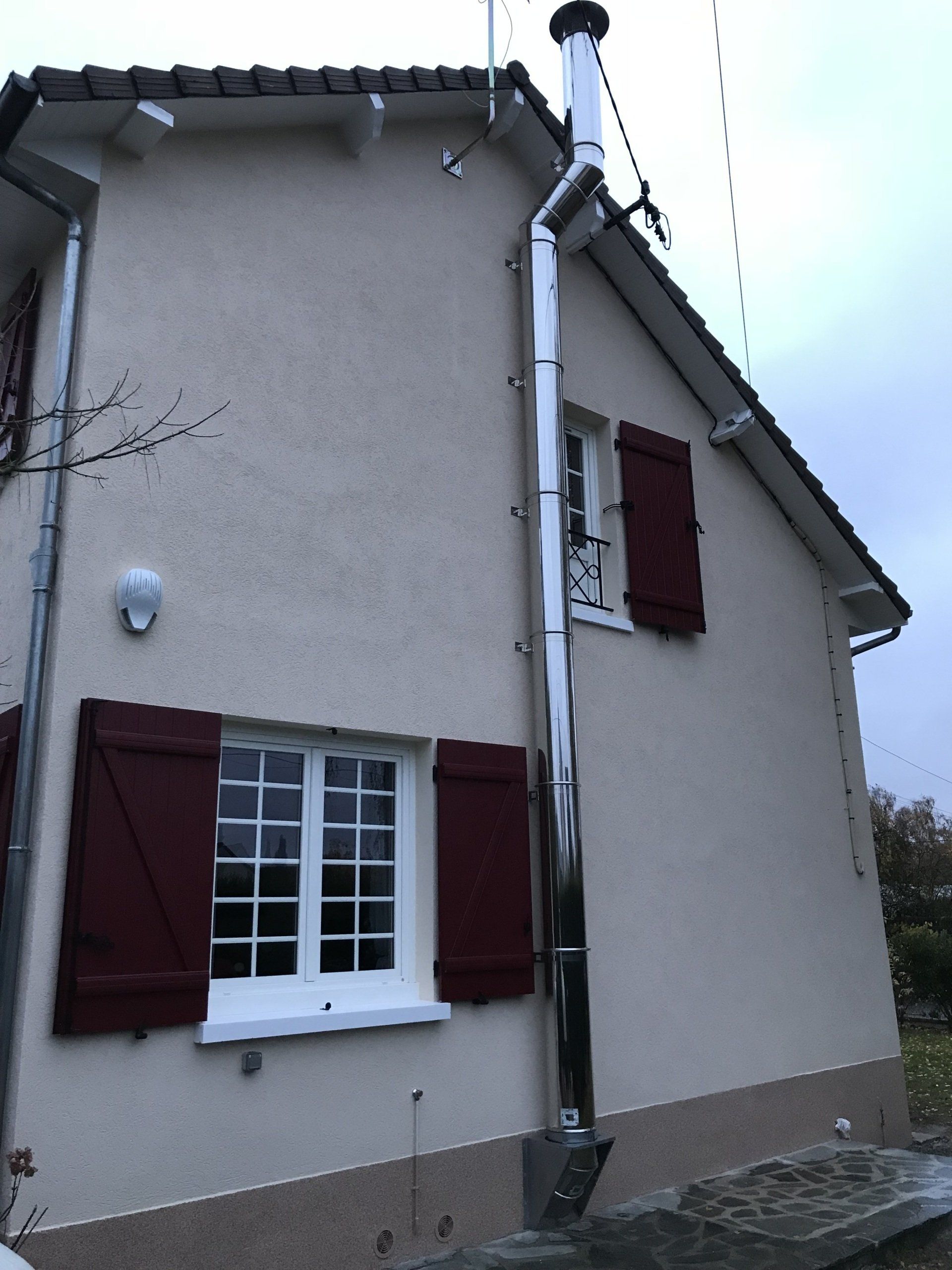Extérieur d'une maison avec une cheminée en acier inoxydable. Murs beiges, volets rouges, ciel nuageux.