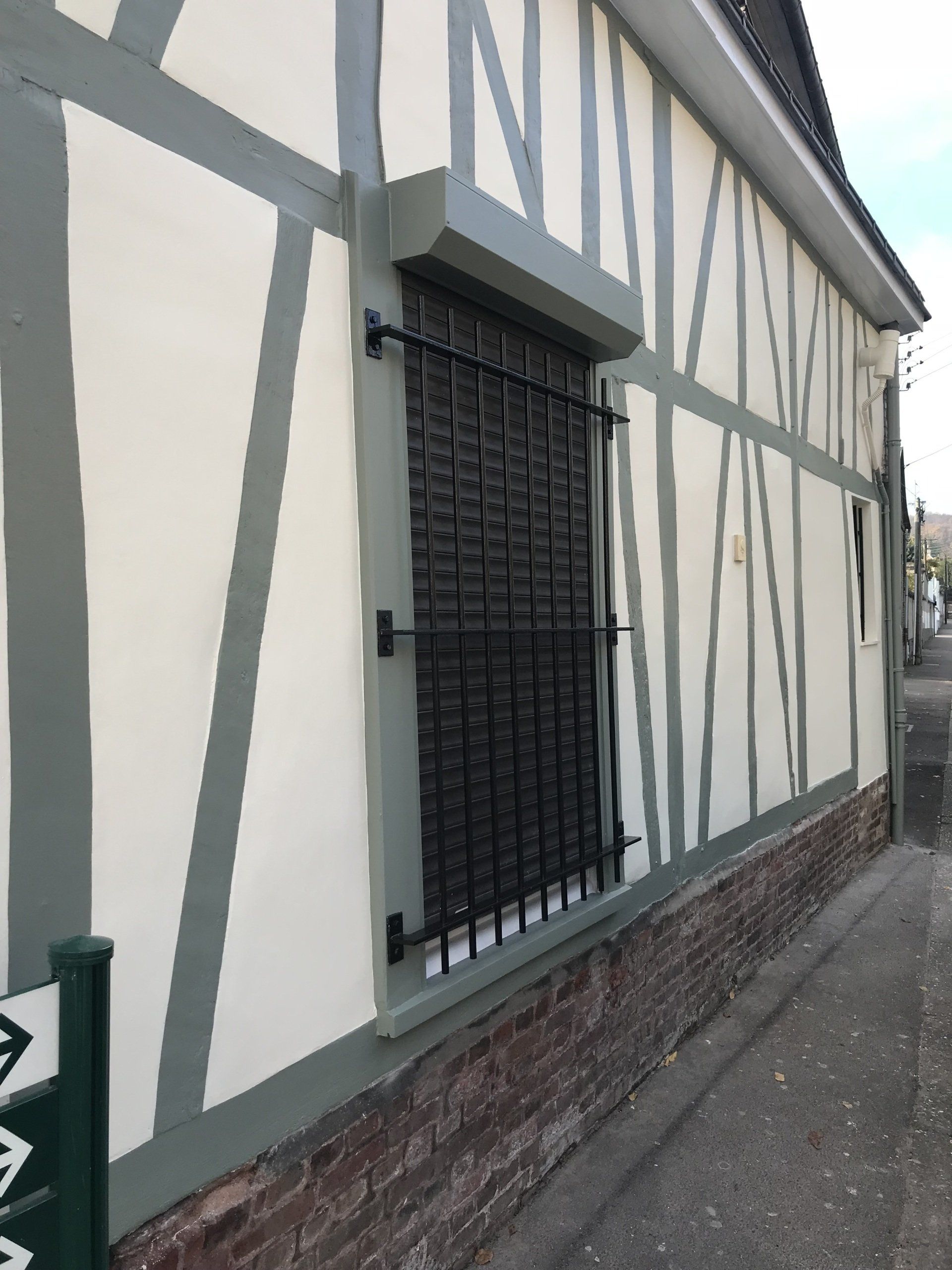Bâtiment à colombages avec une fenêtre protégée par une grille noire. Les boiseries sont grises et le mur est peint en blanc.