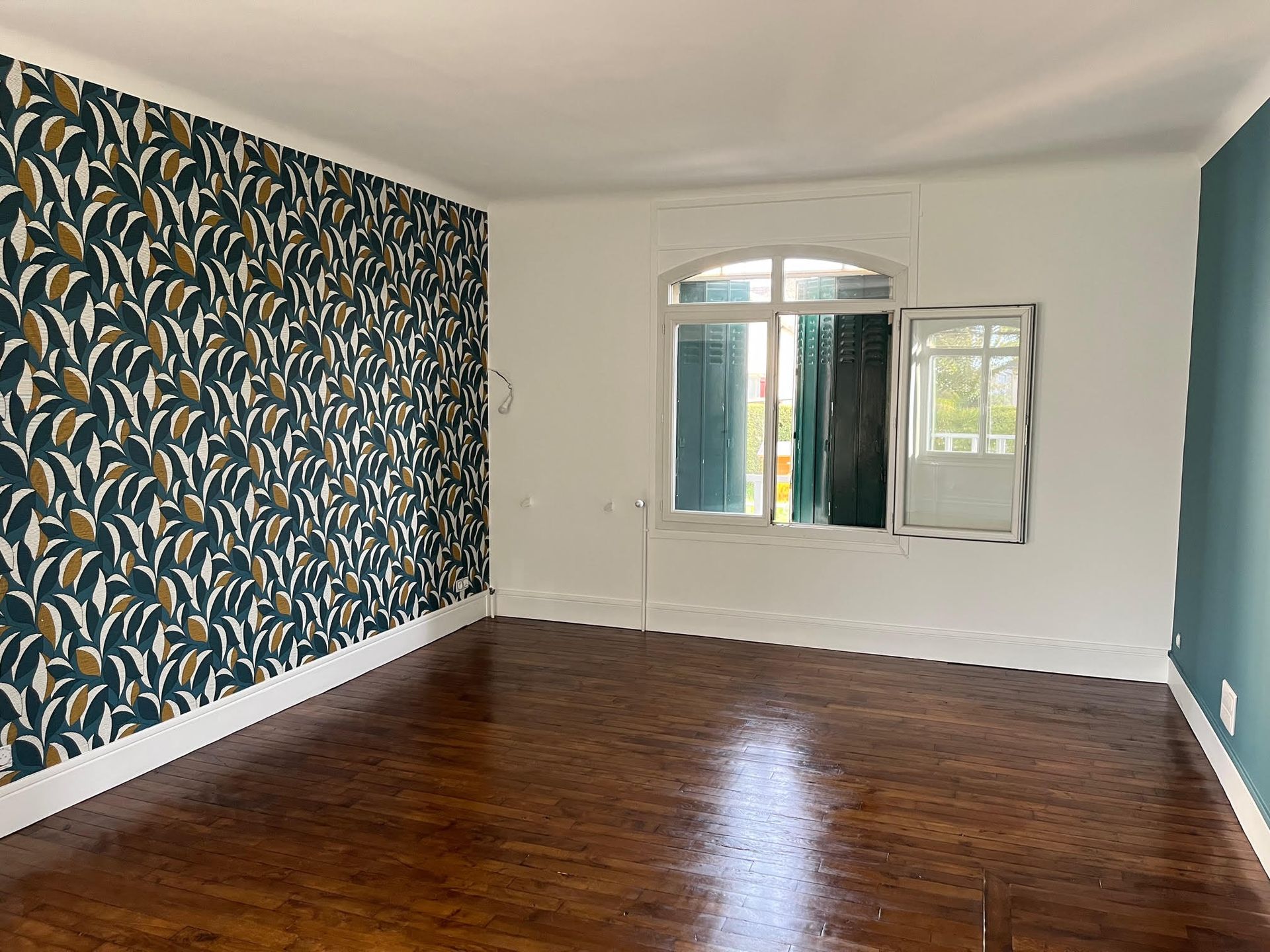 Pièce vide avec papier peint à motifs floraux, mur turquoise, fenêtre cintrée et parquet.