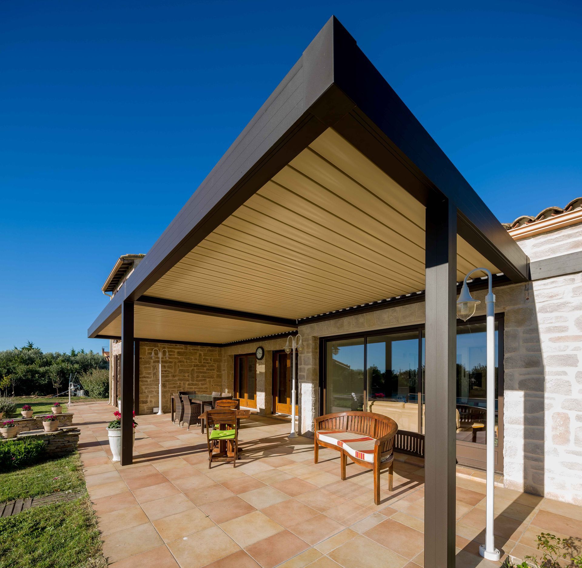 Patio avec cadre sombre, toit beige et mobilier d'extérieur contre une maison en pierre sous un ciel bleu.