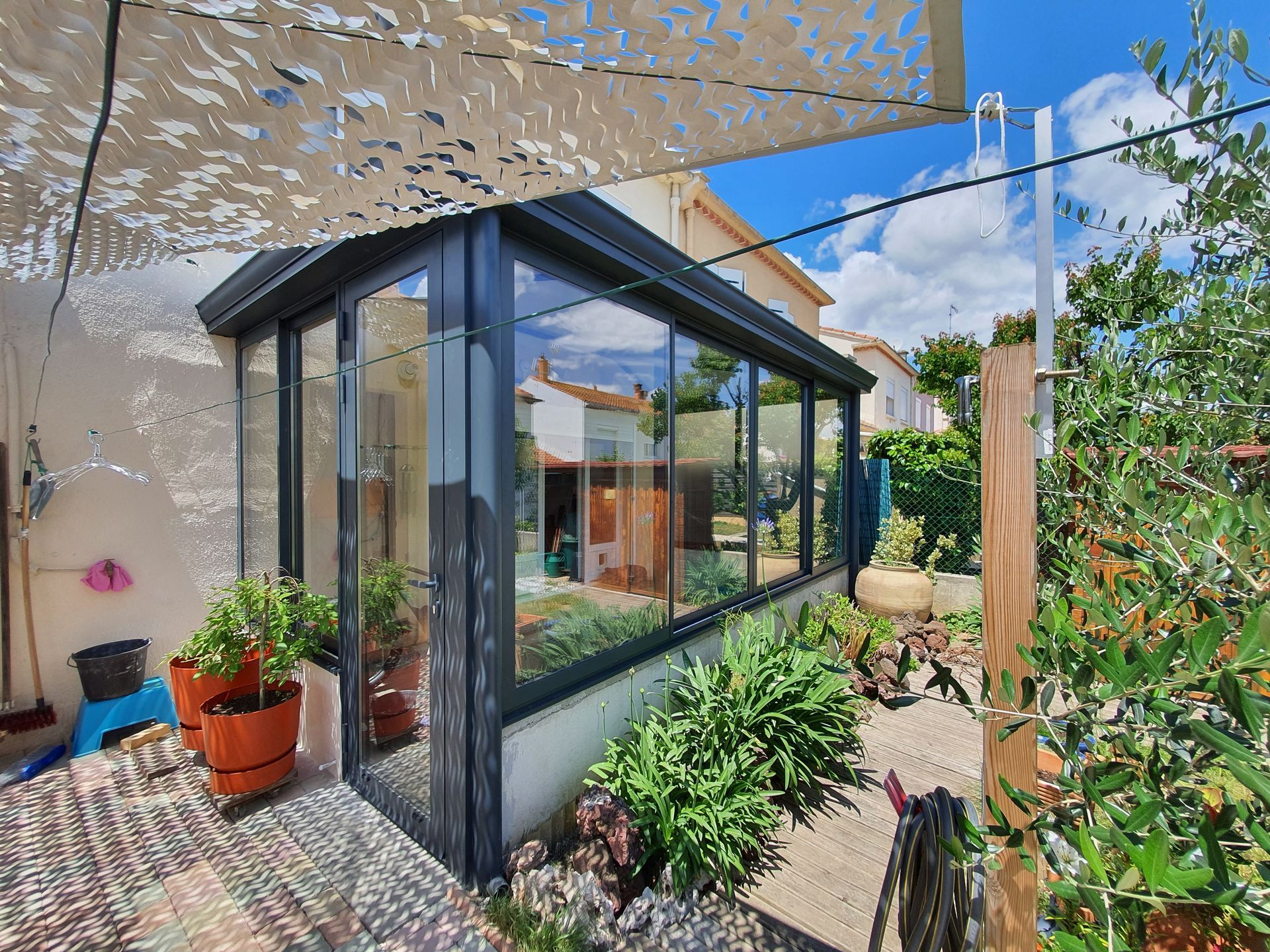 Véranda aux parois vitrées, entourée de plantes, sous un auvent par une journée ensoleillée.