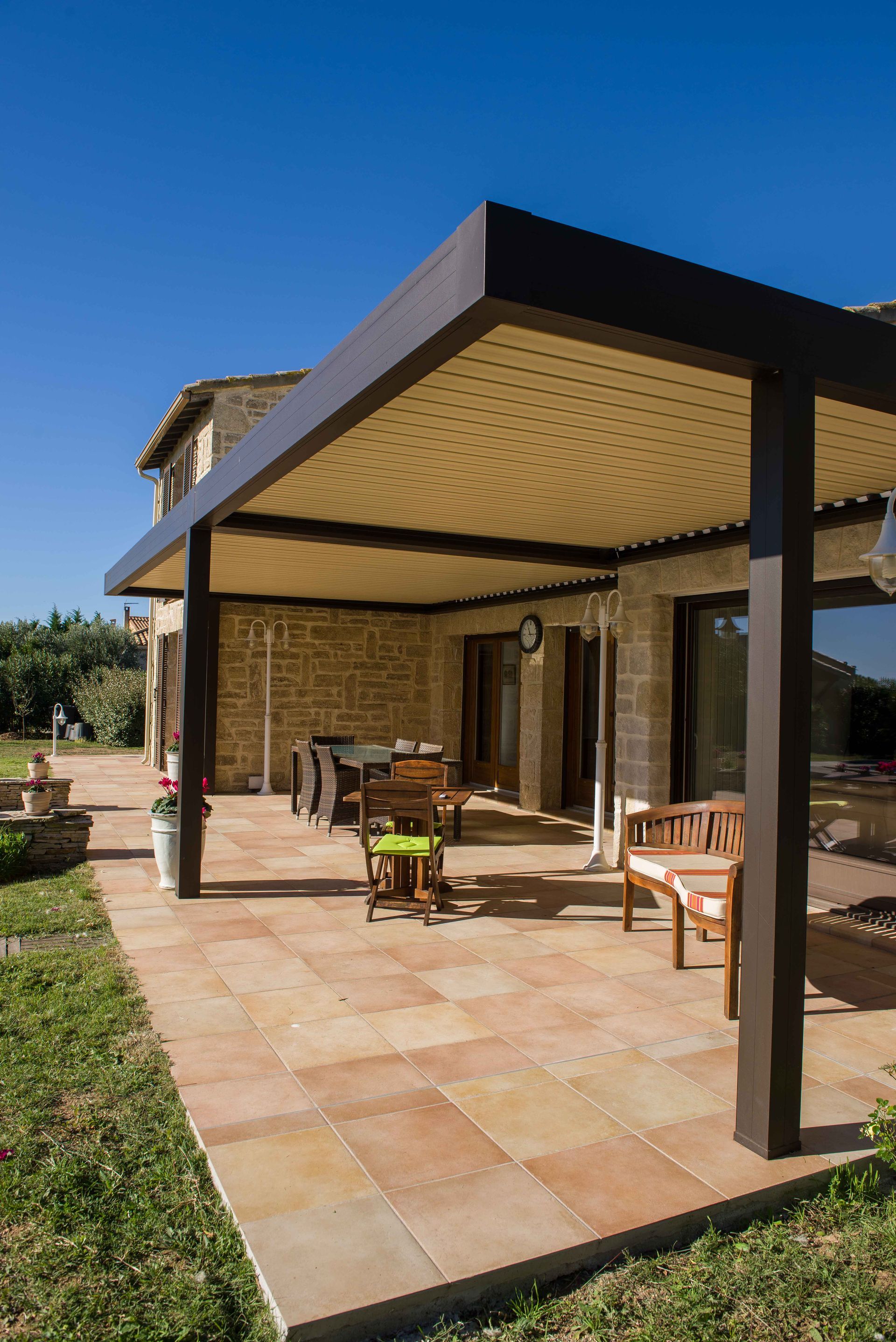 Patio avec pergola marron, sol carrelé, mobilier en bois et maison en pierre sous un ciel bleu.