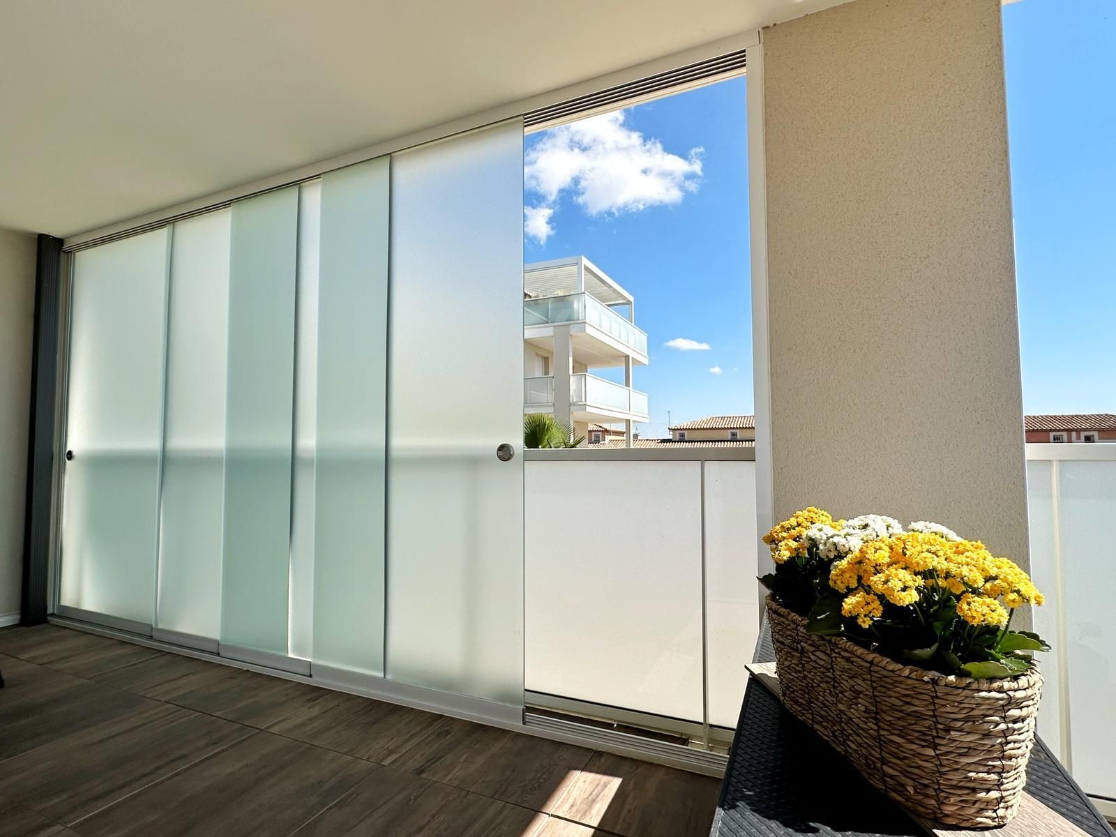Balcon avec portes en verre dépoli, partiellement ouvertes, montrant un ciel bleu vif et des fleurs.