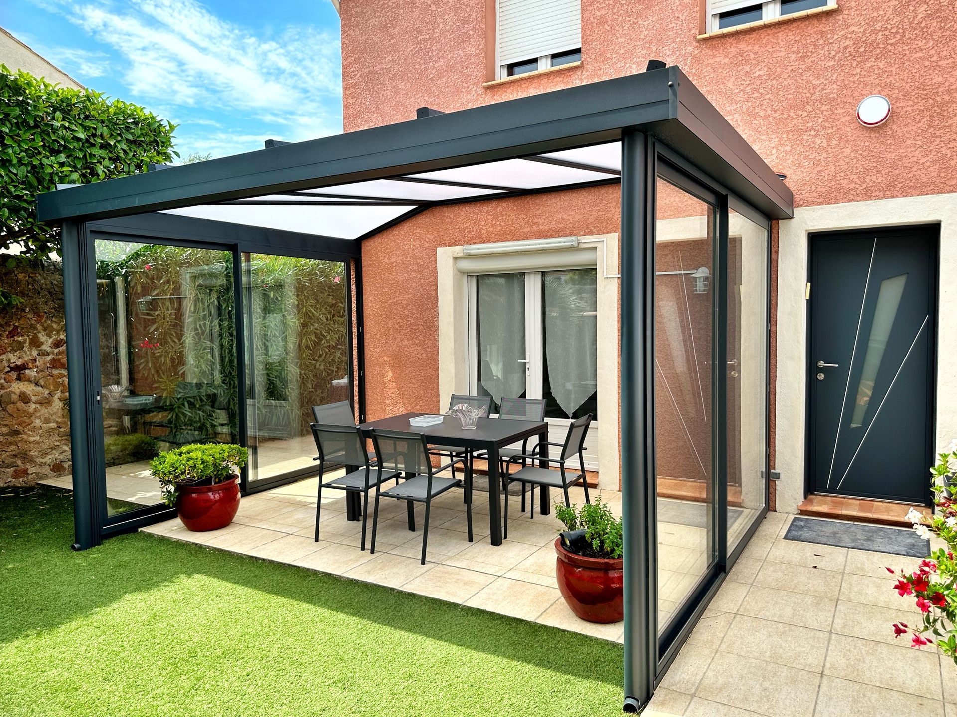 Patio fermé avec parois vitrées et cadre noir, comprenant une table et des chaises sur un patio à côté d'une maison.