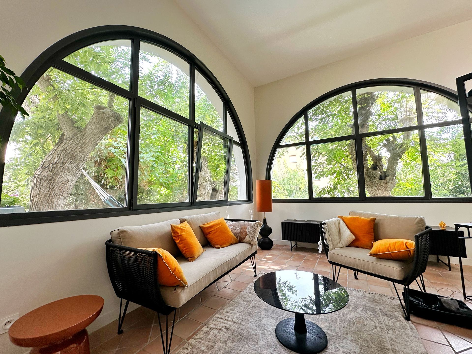 Fenêtres cintrées donnant sur les arbres, comprenant un canapé beige, des coussins orange et une table basse ronde.